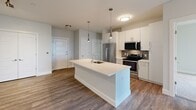 Located at 540 Revere Beach Blvd in Revere, MA, this thoughtfully designed 2-bedroom, 1-bathroom residence offers a clean, contemporary aesthetic across a single floor. The open-concept kitchen and living area serves as the heart of the home, anchored by a large center island with a white quartz countertop, undermount sink, and stylish pendant lighting. White shaker cabinetry, a mosaic tile backsplash, and a full suite of stainless steel appliances—including a refrigerator, range, and microwave—complete the kitchen. Rich hardwood-style flooring flows throughout the main living area, and a window at the far end of the living space fills the room with natural light. The two bedrooms feature plush wall-to-wall carpeting and neutral tones, with the primary bedroom offering comfortable proportions and a large closet. One bedroom benefits from a generous window with outdoor views of greenery and water. The bathroom is finished with wood-look tile flooring, a modern vanity with white countertop, a large mirror, and a tiled walk-in shower, delivering a clean and functional design. Overall, the space presents a bright, modern atmosphere with cohesive finishes throughout.