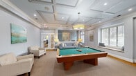 Located in Greer, South Carolina, this well-appointed community recreation room offers a sophisticated and inviting environment designed for leisure and social gatherings. The space is anchored by a full-size billiards table positioned at the center beneath an elegant coffered ceiling with recessed lighting and a stylish drum pendant fixture, lending the room a refined, upscale ambiance.

A built-in wet bar and counter area with light stone countertops, dark wood cabinetry, and bar seating provides a convenient hub for entertaining, complemented by a mounted flat-screen television set into a tastefully designed accent wall with arched niches. Adjacent to the bar, a dedicated lounge area features plush blue sofas and cream armchairs arranged for comfortable conversation, with decorative artwork and warm lamp lighting enhancing the welcoming atmosphere.

The flooring transitions between warm-toned hardwood near the entry and bar zone and soft neutral carpet in the lounge and billiards areas. Large windows dressed with blinds allow natural light to filter through, while a glass-paneled door connects the space to additional amenities beyond. The soft gray walls, tray ceiling details, and carefully curated furnishings work together to create a polished, community-focused retreat that balances function with style.