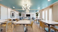 This single-floor communal space is thoughtfully designed with a cohesive coastal-inspired palette, blending teal, cream, and warm wood tones throughout. The dining area is a standout feature, showcasing a striking teal lacquered tray ceiling adorned with two sculptural mid-century modern chandeliers that serve as focal points. Recessed lighting complements the chandeliers, ensuring the space is well-lit throughout. The flooring features a herringbone-patterned light wood tile, adding visual texture and elegance underfoot.

Multiple dining tables are arranged throughout the room, accompanied by a mix of cream, tan, and powder-blue upholstered chairs that reinforce the space's color story. Lush fiddle-leaf fig trees and framed artwork on the walls contribute to the refined yet comfortable ambiance. A mounted flat-screen television is positioned at one end of the room, while sheer curtain-dressed windows allow ample natural light to filter in.

The kitchen area features dark espresso cabinetry, stainless steel appliances, and a spacious island with decorative patterned tile accents and a white quartz countertop. Teal upholstered barstools line the island, tying the kitchen's design seamlessly to the broader space. Wood-look plank flooring extends throughout, unifying the entire area.