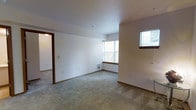 This single-floor residential space offers a functional and straightforward layout designed for comfortable everyday living. The main living area is finished with wall-to-wall carpet in a neutral gray tone, providing a soft and practical underfoot surface that complements the clean, white walls throughout. Warm wood-toned baseboards and door trim add a subtle contrast, lending a touch of traditional character to the otherwise minimalist interior. Natural light enters through multiple windows, including a larger window that draws the eye and brings in views of the surrounding outdoors, as well as a smaller blinds-fitted window that allows for privacy and light control. A wall-mounted heating unit is conveniently positioned beneath the main window, ensuring efficient climate control within the space. An open doorway connects the living area to an adjacent room, enhancing the sense of flow and accessibility between spaces. The ceilings are smooth and standard height, contributing to a bright and open atmosphere. Overall, the space presents a clean, move-in ready environment with a practical layout, neutral finishes, and ample natural light — well suited for a variety of living arrangements.