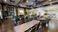 This well-appointed community space serves as a central gathering and dining area, thoughtfully designed to accommodate a large number of residents or guests. The main dining room features multiple rectangular tables paired with comfortable green and brown upholstered chairs, arranged to encourage both intimate and group dining experiences. Overhead, a coffered ceiling with rich dark wood beams adds architectural character, while multiple ceiling fans ensure a comfortable, well-ventilated environment throughout. Recessed lighting and pendant fixtures provide warm, even illumination across the space. A wall-mounted flat-screen television serves as a focal point on one end of the room, complemented by a decorative sideboard and tasteful wall art. An elegant round wall clock and arched decorative accents lend a classic, homey aesthetic to the surroundings. Lush potted plants add touches of greenery throughout. Toward the rear, an adjoining kitchen area is visible, equipped with stainless steel appliances, suggesting full-service meal preparation capabilities. The overall ambiance is warm, inviting, and functional — ideal for communal dining, social events, and everyday gatherings.