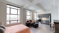 This open-concept studio is thoughtfully designed to maximize functionality within a single, unified living space. Large floor-to-ceiling windows with dark metal frames line the exterior wall, flooding the interior with natural light and offering elevated views of the surrounding cityscape. The windows are fitted with roller shades, providing privacy when needed.

The layout seamlessly integrates the sleeping, living, and kitchen areas. The sleeping zone is positioned near the windows, benefiting from natural light, while a compact seating area with a dark sofa and ottoman serves as a casual living space. Light hardwood flooring runs throughout, contributing to a cohesive and airy aesthetic.

The kitchen is well-appointed with grey cabinetry, stainless steel appliances — including a microwave, stove, and dishwasher — and a light-toned backsplash. Recessed under-cabinet lighting adds both practicality and warmth to the cooking area.

Exposed ceiling beams add an architectural character to the space, complementing the neutral wall tones. Overall, the studio offers a modern, urban ambiance with efficient use of space and strong natural light throughout.
