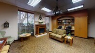 This single-floor community space is thoughtfully designed to offer both relaxation and productivity. The layout flows across several interconnected areas, unified by warm-toned patterned carpeting and a color palette of earthy browns and soft greens. Recessed drop ceilings with flush-mounted skylight panels provide consistent, diffused lighting throughout, complemented by decorative iron chandeliers that add a classic touch.

The lounge area is furnished with floral and patterned upholstered sofas and wingback armchairs arranged around small wooden coffee tables, creating intimate conversation nooks. A decorative fireplace with a wooden mantle serves as a focal point in one section, flanked by large windows that allow natural light and outdoor views. An ornate round mirror with an arched wall surround adds architectural character to the reading room.

Multiple built-in and freestanding bookshelves are filled with a diverse collection of books, reinforcing the library atmosphere. A dedicated computer workstation area features dual desks set within a built-in shelving unit under an arched alcove, providing a functional workspace. Additional cabinetry offers storage throughout. Framed artwork and decorative accents complete the warm, community-centered ambiance.