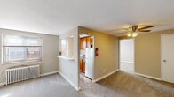 This single-floor residence offers a functional and straightforward layout across several interconnected rooms. The living area is carpeted and opens toward the kitchen via a pass-through window, creating a connected feel between the two spaces. A ceiling fan with a light fixture is positioned overhead, providing both air circulation and ambient lighting. Natural light enters through multiple windows, including one in the living area fitted with a classic cast-iron radiator beneath it, indicating a traditional heating system.

The kitchen is compact and practical, featuring warm-toned wood cabinetry, a white electric range, and a sink with laminate countertops. A window above the stovetop area allows natural light to brighten the workspace. The kitchen is accessible through a dedicated doorway from the main living space.

The bedroom is generously proportioned with plush carpeting and benefits from an abundance of closet space, including multiple reach-in closets with built-in shelving and hanging rods. The entryway to the unit transitions from hard-surface flooring to carpet, clearly delineating arrival from living areas.

The bathroom includes a vanity with storage, a toilet, and a shower-tub combination. Wood-look flooring adds a clean, practical finish underfoot. Overall, the space presents a neutral palette and a practical design throughout.