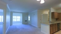 This single-floor residence offers a clean, open layout designed to maximize comfort and functionality. The main living area is finished with plush wall-to-wall carpeting that adds warmth underfoot, while crisp white crown molding along the ceiling provides an elegant architectural detail. Multiple windows allow natural light to fill the space, creating an airy, welcoming ambiance throughout.

A partial wall separates the living area from the kitchen, which features a raised breakfast bar topped with granite countertops — an ideal spot for casual dining or entertaining. The kitchen area is finished with smooth tile flooring, offering a practical contrast to the carpeted living space. Recessed cabinetry and a sleek countertop surface add to the kitchen's functional appeal.

The neutral, light-toned walls throughout the unit provide a versatile backdrop, ready to accommodate a variety of interior styles. A ceiling light fixture in the living area ensures the space is well-lit, complementing the natural light brought in by the windows. The overall ambiance is bright, tidy, and move-in ready, offering a straightforward and livable floor plan suitable for a range of occupants.