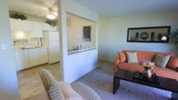 This single-floor residence offers a practical and welcoming layout designed for comfortable everyday living. The living area is carpeted and furnished with warm-toned seating, creating an inviting atmosphere anchored by a dark wood coffee table and a wall-mounted mirror that adds depth to the room. A defining architectural feature is the half-wall pass-through that connects the living space to the kitchen, providing an open feel while maintaining a sense of separation between the two areas. The pass-through ledge serves as a decorative display surface, adding a personalized touch to the layout. The kitchen is finished with white cabinetry and appliances, including a full-size refrigerator, and benefits from a ceiling fan with integrated lighting that brightens the space. Textured ceilings are present throughout, contributing to the space's classic residential character. The neutral wall tones in soft greens and whites create a light, airy ambiance that complements the natural flow between rooms. Overall, the space is thoughtfully arranged to balance openness and function, making it an efficient and comfortable single-level home.