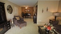 This single-story space features an open-concept layout that seamlessly connects the living and dining areas, creating a cohesive and functional environment. The living area is anchored by plush, dark-upholstered seating, including an armchair with an ottoman, positioned over soft wall-to-wall carpeting that runs throughout the space. Warm beige walls contribute to the inviting ambiance, complemented by decorative wall sconces and tasteful accents.

The dining area, situated toward the rear of the space, accommodates a dark wood dining table with matching chairs, centered beneath a ceiling fan with an integrated light fixture that provides overhead illumination. A framed piece of wall art enhances the dining area backdrop, while a potted plant adds a natural element.

A dark wood storage cabinet or credenza serves as a functional divider between the two areas, offering both storage and display space. The overall aesthetic is cohesive and neutral, with warm earth tones throughout. An entry door is visible along one wall, and the layout provides a practical flow between the primary living zones, making the space feel both organized and welcoming.