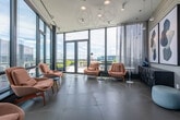 This elegantly designed upper-floor space offers a seamless blend of modern architecture and refined interior styling. The layout consists of a spacious lounge area and a connecting corridor, both unified by expansive floor-to-ceiling curtain wall windows framed in dark steel, which flood the interiors with abundant natural light and frame sweeping panoramic views of the surrounding cityscape and waterfront beyond.

The lounge area is appointed with a curated arrangement of plush terracotta-toned armchairs accented by mid-century modern legs, complemented by low-profile round side tables in muted blue and white tones. Sculptural pendant light fixtures with an open circular wire design hang overhead, adding an artistic focal point against the high ceilings. Sheer curtains line portions of the glazed walls, softening the light while preserving the open, airy atmosphere. Glass doors lead out to an exterior terrace, further extending the connection to the outdoors.

The gallery-style corridor runs alongside the full-length window wall, showcasing vibrant large-scale artwork displayed on a clean white wall, creating an immersive visual experience as one moves through the space. Subtle pendant lighting illuminates the passage, enhancing the art gallery feel. Additional seating alcoves are tucked along the windows, offering intimate vantage points overlooking the waterfront. The interior transitions are finished with textured wall treatments and neutral large-format tile flooring throughout, contributing to the space's polished, contemporary character.