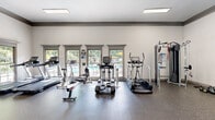 This fitness center offers a comprehensive workout environment housed within a single, open floor plan. The space is designed with functionality and comfort in mind, featuring durable speckled rubber flooring throughout that provides both safety and resilience underfoot. Elevated ceilings lend an airy, spacious feel, while large, framed windows along one wall flood the interior with natural light and provide views of an outdoor pool area, creating an inviting atmosphere for exercise.

The cardio zone is well-appointed with multiple treadmills, elliptical trainers, and a stationary bike, all positioned to take advantage of the poolside views. A dedicated strength training area features a Smith machine with an adjustable bench and a full rack of dumbbells, complemented by a cable and resistance machine station. Full-length mirrors line portions of the walls, enhancing the sense of space and allowing users to monitor their form. Wall-mounted televisions are positioned throughout for entertainment during workouts.

The interior is finished in a clean, neutral color palette of white and soft gray, with subtle blue accent walls adding a modern touch. Recessed and flush-mounted ceiling lights ensure consistent illumination. French doors connect the fitness center to an adjacent common area, reinforcing its role as a shared amenity within a larger facility.