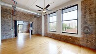 This urban loft seamlessly blends industrial character with modern finishes across a single, well-appointed floor. The open living area makes a striking first impression with its exposed brick walls, soaring ceilings featuring visible ductwork and pipes, and light hardwood flooring that flows throughout. Large black-framed windows flood the space with natural light and frame expansive city views, while a ceiling fan and rail lighting add functional style overhead.

The kitchen is thoughtfully equipped with rich wood cabinetry, granite countertops, stainless steel appliances including a refrigerator, microwave, and double wall oven, complemented by a door that opens directly to an outdoor area. Exposed brick continues here, reinforcing the loft's cohesive aesthetic.

Two hallways connect the living spaces to the bedrooms, one featuring a structural column and track lighting. Both bedrooms are carpeted with soaring ceilings, offering a calm retreat from the open living areas. The bedrooms include generous closet space with warm wood doors.

The two bathrooms are finished to a high standard. The primary bath features a glass-enclosed walk-in shower with white subway tile, dark accent trim, and a granite-topped vanity. The second bathroom offers a tub-shower combo with floor-to-ceiling tile, a wood-paneled sliding door, and a matching granite vanity.