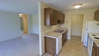 This single-floor residence offers a practical and functional layout designed for comfortable everyday living. The kitchen is a central feature of the home, outfitted with rich dark wood cabinetry that provides ample upper and lower storage space. A double-basin sink sits beneath the cabinetry along a clean white countertop, complemented by a dishwasher and additional lower cabinet storage. The kitchen floor is finished with classic tile, adding durability to a high-traffic area. A structural column subtly defines the boundary between the kitchen and the adjoining living space, maintaining an open and connected feel throughout. The living area features wall-to-wall carpeting, lending a warm and comfortable atmosphere. Textured ceilings are consistent throughout the home, and flush-mount lighting provides overhead illumination. Neutral wall tones create a versatile backdrop, allowing for a variety of interior styles. Doorways visible from the main areas hint at additional rooms beyond, suggesting a well-organized floor plan. Overall, the space presents a straightforward, move-in-ready environment with classic residential finishes throughout.