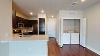 This single-story residence offers a thoughtfully designed open-concept layout where the kitchen flows seamlessly into the living area. Rich, dark hardwood-style flooring runs throughout the main living spaces, creating a warm contrast against the neutral-toned walls and bright white trim.

The kitchen serves as a central hub, anchored by a peninsula island with a light quartz countertop, an undermount sink, and a stainless steel dishwasher. Dark espresso cabinetry lines the kitchen walls, complemented by stainless steel appliances including a refrigerator, microwave, and range. Recessed lighting illuminates the workspace effectively.

The living area is bright and airy, benefiting from a large sliding glass door dressed with vertical blinds that invites natural light and outdoor views. A ceiling fan overhead adds both function and comfort to the space.

A dedicated in-unit laundry alcove, conveniently located off the kitchen, houses a full-size washer and dryer. The primary bathroom features a dark vanity with quartz countertops, a large mirror, and direct access to a carpeted walk-in closet equipped with built-in shelving and hanging rods for ample storage.