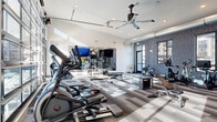 This fitness center at 5025 W 20th St in Greeley, Colorado, is a thoughtfully designed exercise space that combines modern architecture with a full suite of workout amenities. The single-floor layout makes efficient use of its generous footprint, creating an open, airy environment ideal for a variety of training styles.

The room features soaring ceilings that amplify the sense of space, complemented by an oversized glass garage door along one wall that floods the interior with natural light and offers views of the surrounding exterior. Clerestory windows along the upper walls further enhance brightness throughout the day. A large ceiling fan keeps air circulating, and exposed ductwork adds an industrial-modern aesthetic touch.

The exercise area is comprehensively outfitted with cardio equipment including treadmills, ellipticals, and stationary bikes positioned to take advantage of the natural light. A dedicated strength training zone features a cable/squat rack, adjustable benches, a full dumbbell rack, and a plyo box for functional training. Rowing machines round out the cardio offerings.

Floor-to-ceiling mirrors along one wall visually expand the space and support form-monitoring during workouts. A bold, patterned accent wall adds a dynamic visual element, while the multi-toned striped carpet tiles provide both durability and contemporary style. Wall-mounted flat-screen TVs offer entertainment throughout the workout. The space is bright, clean, and well-organized for an effective fitness experience.