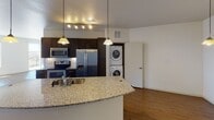 This contemporary single-floor residence offers a thoughtfully designed layout with clean lines and a bright, open atmosphere throughout. The spacious main living area is anchored by rich hardwood-style flooring and flooded with natural light through large sliding glass doors that open to a private balcony, offering sweeping elevated views of the surrounding cityscape and beyond.

The kitchen is a standout feature, showcasing dark espresso cabinetry paired with granite countertops, a curved breakfast bar peninsula, and a full suite of stainless steel appliances including a range, over-the-range microwave, and French door refrigerator. A tile backsplash and pendant lighting add refined detail, while a stacked in-unit washer and dryer provide everyday convenience.

The bedrooms are carpeted for comfort and benefit from generous window placement that continues the theme of elevated outdoor views and ample natural light. Neutral white walls throughout create a versatile, move-in-ready canvas. The bathroom, visible from the living area, features warm-toned finishes. The overall design balances modern aesthetics with functional living, making this a well-rounded and inviting space suited for a variety of lifestyles.