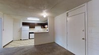 This single-floor unit presents a compact, open-concept layout where the living area flows directly into the kitchen, creating a connected and functional living environment. The living space features wall-to-wall carpeting and painted concrete block walls, giving the space a sturdy, utilitarian character. A textured ceiling with flush-mounted lighting provides even illumination throughout. The kitchen is delineated by a raised peninsula countertop that serves as a natural dividing element between the two spaces. It is equipped with dark espresso-toned cabinetry, a white refrigerator, a white range, and a built-in dishwasher. A single-basin sink is positioned along the peninsula with overhead pendant lighting above. The kitchen flooring transitions to a checkered vinyl tile, providing a practical and easy-to-maintain surface. A white panel door leads out from the main living area, offering access to additional spaces. Overall, the unit is designed with practicality in mind, making efficient use of its footprint while maintaining a cohesive flow between the kitchen and living areas.