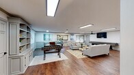 Located at 201 NC-54 W in Carrboro, North Carolina, this single-floor community space offers a generously sized and thoughtfully designed layout ideal for residents to gather and relax. The open-concept living room is a standout feature, boasting rich hardwood flooring, wainscoting along the lower walls, and flush panel lighting that keeps the space bright and inviting. A foosball table and shuffleboard table provide recreational options, while a wall-mounted flat-screen TV and ceiling fan add comfort and entertainment. A seating arrangement with neutral-toned sofas and armchairs anchors the lounge area, complemented by decorative artwork and natural light flowing in through windows dressed with woven Roman shades. Built-in shelving with cabinetry adds both function and charm. The dining area offers a dedicated space for communal meals, with ample natural light from surrounding windows. The entrance features wainscoted walls, warm hardwood floors, and a clean, welcoming aesthetic. A well-equipped laundry room provides multiple side-by-side washers and stacked dryer units, a central folding table, and wainscot detailing consistent with the rest of the space. Throughout, the design maintains a cohesive, neutral palette that is both practical and polished.