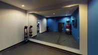 This fitness center is a thoughtfully designed, single-floor facility built to support a wide range of workout routines. The space is divided into distinct functional zones, each tailored to specific training needs. Bold blue accent walls contrast with neutral gray surfaces throughout, creating an energetic and motivating atmosphere. Recessed ceiling lighting provides consistent, even illumination across all areas.

The free weight and functional training zone features rubber flooring for safety and durability, and is equipped with a dumbbell rack, medicine ball rack, stability balls, and a wall-mounted flat-screen television for entertainment or instructional use. A full-wall mirror spans one side of this area, enhancing the sense of openness and allowing users to monitor their form during workouts.

An adjacent cardio area is visible through a connecting doorway and houses a row of treadmills alongside additional strength training equipment, offering ample space for cardiovascular exercise. The rubber flooring continues throughout, ensuring a consistent and functional surface underfoot.

The overall design of the facility emphasizes practicality and performance, with a clean, modern aesthetic reinforced by the coordinated color palette and organized equipment layout. The space is well-suited for individual training sessions across a broad spectrum of fitness goals.