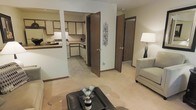 This single-floor residential space offers a welcoming and functional layout designed for comfortable everyday living. The open-concept living area serves as the heart of the home, featuring neutral-toned carpeting, a stylish dark leather coffee table, and warm ambient lighting provided by a classic floor lamp. A plush seating area creates an inviting atmosphere, ideal for relaxing or entertaining. Decorative wall accents add a subtle touch of personality to the space.

Adjacent to the living area, the kitchen is efficiently laid out with ample cabinetry in a warm wood finish paired with light-colored countertops. The kitchen is equipped with essential appliances, including a range and dishwasher, and benefits from clean, practical tile flooring that contrasts neatly with the carpeted living spaces. The open sightline between the kitchen and living area enhances the sense of openness throughout.

Multiple interior doors with rich, dark wood finishes lead to additional rooms, offering privacy and a well-organized flow throughout the unit. The neutral wall tones and consistent warm color palette throughout the space create a cohesive and calming ambiance. Overall, this thoughtfully arranged single-level space balances practicality with comfort.