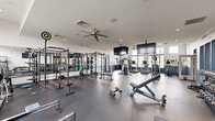 Located at 2100 Haddonfield Road in Pennsauken, NJ, this fitness facility is organized across two dedicated exercise rooms, together comprising a generously sized single-floor layout designed to support a full spectrum of workout routines.

The main exercise room features durable rubber flooring and is outfitted with an impressive array of strength and cardio equipment, including power racks with weight plates, adjustable benches, dumbbells, kettlebells, medicine balls, resistance equipment, and a full rack of free weights. Multiple Matrix-brand treadmills and elliptical machines line the perimeter, while wall-mounted flat-screen televisions and ceiling fans enhance the workout environment. Large windows along the upper walls allow natural light to fill the space, and mirrored walls help expand the sense of openness. A motivational wall decal adds an energizing touch to the atmosphere.

The second exercise room serves as a dedicated cycling studio, finished with light wood-look tile flooring and warm neutral-toned walls. It is equipped with a row of interactive stationary bikes featuring individual display screens, ideal for on-demand or instructor-led cycling sessions. Wall mirrors and large windows with roller shades provide a bright, focused environment. The two rooms are connected, offering a seamless transition between workout disciplines within one cohesive fitness facility.