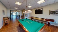This single-floor recreational space is designed to serve as a social and entertainment hub. At its center sits a full-size pool table with a classic wooden frame and green felt surface, illuminated by a stylish pendant light fixture suspended from a drop-panel ceiling. Alongside the pool table, a shuffleboard table offers additional gaming options, while a large wall-mounted flat-screen TV provides a focal point for viewing entertainment.

The room features warm hardwood flooring throughout, complemented by neutral-toned walls that create a welcoming ambiance. A wall-mounted cue rack keeps equipment neatly organized and within reach. Recessed and panel lighting ensure the space is well-lit for gameplay and socializing.

An adjacent area visible through the room connects to a bar-height seating section, expanding the social capacity of the space. Large floor-to-ceiling dark-framed windows and a glass door open directly onto a spacious outdoor patio, flooding the interior with natural light and offering views of a beautifully appointed exterior courtyard. The patio features stone tile flooring, decorative seating, and is framed by stone-accented architectural columns, enhancing the overall aesthetic of the space and creating a seamless indoor-outdoor experience.