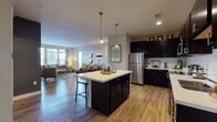 This single-floor residence offers a thoughtfully designed open-concept layout that seamlessly connects the kitchen, dining, and living areas. The kitchen serves as a central hub, outfitted with dark espresso cabinetry extending to the ceiling, white subway tile backsplash, stainless steel appliances, and light-toned countertops. A large central island with seating provides additional workspace and casual dining. Pendant lighting overhead adds a refined touch, while wood-style flooring flows throughout the main living spaces.

The open living and dining area benefits from natural light streaming through large windows dressed with curtains, creating a bright and welcoming atmosphere. The dining nook features hardwood-style flooring and elegant wall accents.

The unit includes two bedrooms, each carpeted and featuring windows with blinds and curtain panels. One bedroom is appointed with warm neutral tones, while the second features deeper, moodier hues with teal accents for a distinct character.

The bathroom is finished with dark vanity cabinetry, white countertops, a large mirror with lighted fixtures, and a tiled tub-shower combination, maintaining the cohesive design palette found throughout the residence.