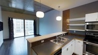 This single-floor space showcases a thoughtfully designed open-concept layout that blends industrial and modern aesthetics. The kitchen serves as a central feature, anchored by a substantial peninsula with a dark countertop and a stainless steel double-basin sink. White cabinetry below the counter provides clean contrast against the darker tones throughout the space. A striking mosaic tile backsplash in warm copper and brown tones adds texture and visual depth, complemented by open white shelving mounted against a warm-toned accent wall — a practical yet decorative storage solution. Two round, globe-style pendant lights hang from above, casting a warm, ambient glow over the workspace. The ceiling incorporates exposed concrete paneling, reinforcing the industrial character of the space. Dark hardwood flooring runs throughout, grounding the open layout with a rich, cohesive tone. A large window at the far end of the space floods the interior with natural light and frames an urban cityscape, offering views of surrounding multi-story buildings. The overall ambiance is contemporary and urban, with a balance of raw materials and refined finishes.