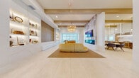 Located at 1799 N Williamson Blvd in Daytona Beach, Florida, this single-floor amenity space offers a thoughtfully designed environment for socializing, dining, and productive work. The layout spans a generous footprint and is connected by a series of bright, airy hallways finished in large-format porcelain tile.

The central living room serves as the heart of the space, anchored by a sculptural golden curved sofa beneath a tiered gold chandelier. Warm wood ceiling beams add architectural character, while a built-in illuminated display wall and a wall-mounted fireplace with a marble surround create a refined, welcoming ambiance.

Adjacent to the lounge, a dining area features floor-to-ceiling windows that flood the space with natural light and offer views of the outdoor grounds, including a palm-lined pool area. Natural wood communal tables and bench seating complement the airy aesthetic. A nearby wet bar with dramatic marble backsplash and countertops, backlit shelving, and pendant globe lighting provides an elegant social hub with counter seating.

The office suite includes a comfortable lounge with plush blue accent chairs, a private phone booth, and a glass-enclosed conference room equipped with a large work table and rolling chairs — ideal for focused work or meetings. Throughout, the design palette of warm whites, natural wood, and soft blues creates a cohesive, modern coastal atmosphere.