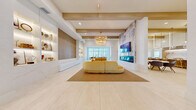 Located at 1799 N Williamson Blvd in Daytona Beach, Florida, this single-floor amenity space offers a thoughtfully designed environment for socializing, dining, and productive work. The layout spans a generous footprint and is connected by a series of bright, airy hallways finished in large-format porcelain tile.

The central living room serves as the heart of the space, anchored by a sculptural golden curved sofa beneath a tiered gold chandelier. Warm wood ceiling beams add architectural character, while a built-in illuminated display wall and a wall-mounted fireplace with a marble surround create a refined, welcoming ambiance.

Adjacent to the lounge, a dining area features floor-to-ceiling windows that flood the space with natural light and offer views of the outdoor grounds, including a palm-lined pool area. Natural wood communal tables and bench seating complement the airy aesthetic. A nearby wet bar with dramatic marble backsplash and countertops, backlit shelving, and pendant globe lighting provides an elegant social hub with counter seating.

The office suite includes a comfortable lounge with plush blue accent chairs, a private phone booth, and a glass-enclosed conference room equipped with a large work table and rolling chairs — ideal for focused work or meetings. Throughout, the design palette of warm whites, natural wood, and soft blues creates a cohesive, modern coastal atmosphere.
