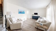 Located in Virginia Beach, VA, this two-story home offers a clean, coastal-inspired design across three bedrooms and two bathrooms. The main floor features light hardwood-style flooring throughout the living, dining, and kitchen areas. The living room is bright and airy, with natural light streaming through large windows dressed in sheer curtains, creating a welcoming and relaxed atmosphere. The dining area is a standout space, highlighted by a chandelier overhead and sliding glass doors that open directly to a private outdoor patio, seamlessly blending indoor and outdoor living. The kitchen is efficiently laid out with ample white cabinetry, granite-look countertops, and full appliances including a range, dishwasher, and refrigerator. A full bathroom with a shower-tub combination is also found on the main level. Upstairs, the primary bedroom offers plush carpeting, natural light through shuttered windows, and a serene, restful ambiance. The additional bedrooms provide comfortable accommodations, including one fitted with a bunk bed and window views. A second full bathroom with a shower serves the upper floor. A closet and hallway complete the upper level. Throughout the home, a neutral white palette and coastal accents create a cohesive, inviting feel.
