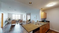 This single-floor residence is designed with an open-concept layout that seamlessly connects the kitchen, dining, and living areas into one cohesive space. The kitchen is well-appointed with warm honey-toned cabinetry, granite countertops, a double stainless steel sink, and stainless steel appliances including a range, microwave, and refrigerator. A breakfast bar peninsula separates the kitchen from the dining area, which comfortably accommodates a round dining table with upholstered chairs. Rich hardwood-style flooring extends throughout the main living areas, adding warmth and continuity to the open floor plan. The living room is bright and inviting, anchored by large windows that allow generous natural light and provide outdoor views. Two bedrooms offer comfortable retreats, each featuring soft carpet, neutral walls, and windows that continue the theme of natural light throughout the unit. The bathroom is finished with tile flooring, warm wood vanity cabinetry, a countertop with an integrated sink, and a walk-in shower enclosed by sliding frosted glass doors. The overall ambiance is clean, modern, and welcoming, with a neutral palette that creates a versatile and functional living environment.
