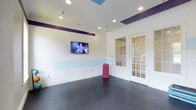 The yoga and fitness studio at Eagle Rock at Freehold is a thoughtfully designed single-floor space that offers residents a dedicated area for exercise and wellness. The room features a clean, open layout with durable dark speckled flooring that is well-suited for yoga, stretching, and light fitness activities. The walls are painted in a crisp white with distinctive purple accent bands and a soft blue horizontal stripe, creating an energizing yet calming atmosphere. Recessed lighting throughout the ceiling provides bright, even illumination across the entire space. A wall-mounted flat-screen television is positioned for easy viewing during workouts, adding a modern, functional element to the room. Decorative tray ceiling details highlighted in purple and sky blue add visual interest overhead. Elegant white French doors with glass pane inserts offer a connection to the adjacent areas of the facility, allowing natural light to filter into the room while maintaining a sense of openness. The overall ambiance is clean, vibrant, and purpose-built to support an active lifestyle, reflecting the community-focused amenities available at Eagle Rock at Freehold.
