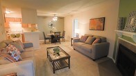 This single-floor space showcases an open-concept design that seamlessly connects the living and dining areas, creating a cohesive and functional layout. The living area is anchored by plush wall-to-wall carpeting in a warm neutral tone, providing a comfortable foundation for the seating arrangement. A glass-topped coffee table with a lower shelf adds a practical yet stylish centerpiece to the space.

The dining area is positioned adjacent to the living room and features a round table with dark wood chairs, complemented by a decorative wall fixture that adds an artistic touch. A ceiling fan with integrated lighting serves the dining zone, ensuring both comfort and illumination. The transition between the two areas flows naturally, making the combined space feel open and inviting.

A pass-through or counter connects to what appears to be a kitchen, offering convenient accessibility between the cooking and living spaces. Warm ambient lighting throughout enhances the cozy atmosphere, while a doorway allows natural light to filter in. Decorative accents, including framed artwork and accent pillows, add personality and warmth to the overall aesthetic.