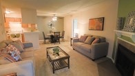 This single-floor space showcases an open-concept design that seamlessly connects the living and dining areas, creating a cohesive and functional layout. The living area is anchored by plush wall-to-wall carpeting in a warm neutral tone, providing a comfortable foundation for the seating arrangement. A glass-topped coffee table with a lower shelf adds a practical yet stylish centerpiece to the space.

The dining area is positioned adjacent to the living room and features a round table with dark wood chairs, complemented by a decorative wall fixture that adds an artistic touch. A ceiling fan with integrated lighting serves the dining zone, ensuring both comfort and illumination. The transition between the two areas flows naturally, making the combined space feel open and inviting.

A pass-through or counter connects to what appears to be a kitchen, offering convenient accessibility between the cooking and living spaces. Warm ambient lighting throughout enhances the cozy atmosphere, while a doorway allows natural light to filter in. Decorative accents, including framed artwork and accent pillows, add personality and warmth to the overall aesthetic.