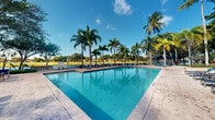 Located at 5555 North Military Trail in Boca Raton, Florida, this outdoor pool area offers a quintessential South Florida experience. The centerpiece is a generously sized swimming pool with vivid, crystal-clear turquoise water that reflects the surrounding palm trees and bright blue sky. The pool's clean geometric shape is complemented by a broad travertine-style deck that provides ample space for lounging, lined with rows of chaise lounge chairs positioned to take in the sun and scenery. Towering royal palm trees frame the area on all sides, lending a lush, resort-like atmosphere. The setting opens to expansive green grounds beyond the pool perimeter, offering serene views of manicured lawns and landscaped surroundings. The combination of abundant natural light, tropical foliage, and open sky creates a bright, airy ambiance ideal for relaxation and recreation. The overall environment evokes the feel of a well-appointed community or resort amenity, blending natural Florida beauty with thoughtfully maintained outdoor infrastructure. This pool area stands as a standout feature of the property, providing an inviting and visually striking outdoor retreat in the heart of Boca Raton.