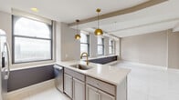 Located at 50 W. Front Street in the heart of downtown Columbus, Ohio, this well-appointed 2-bedroom, 2-bathroom residence offers a refined urban living experience. The single-floor layout is thoughtfully designed, connecting a bright, open-concept living, dining, and kitchen area that serves as the centerpiece of the home. The kitchen is outfitted with stainless steel appliances, a gas range, quartz countertops, gray shaker-style cabinetry, a central island with a double sink, and striking gold globe pendant lights — all complemented by large arched windows that frame sweeping city views. Polished marble-style porcelain tile flows throughout the main living areas and hallway, lending a clean, luminous quality to the space. Both bedrooms feature plush carpeting and windows with urban vistas, with the primary bedroom offering generous proportions and dedicated closet space. The primary bathroom is a standout, showcasing a glass-enclosed walk-in shower with marble tile surround, a soaking tub, and a gray vanity with gold-toned fixtures. A second full bathroom, hallway, two closets, and in-unit laundry complete the functional floor plan. The overall aesthetic is modern and neutral, with warm taupe tones and crisp white trim throughout.