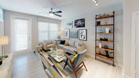 This well-designed single-floor residence offers a cohesive, contemporary aesthetic throughout. The kitchen serves as a focal point, showcasing crisp white shaker cabinetry, light quartz countertops, and a full suite of stainless steel appliances including a French door refrigerator, range, microwave, and dishwasher. A central island with bar seating and pendant lighting above creates a natural gathering space, while light-toned hardwood-style flooring ties the kitchen seamlessly into the adjoining living area.

The living room is bright and welcoming, benefiting from natural light through large windows with blinds and a ceiling fan overhead. The space accommodates a comfortable seating arrangement and features open shelving along one wall for display and storage.

The primary bedroom offers plush carpeting, a ceiling fan, a walk-in closet with built-in organizers, and direct access to the bathroom. A second bedroom provides natural light through dual windows, soft carpeting, and generous closet space. The bathroom features a full tub-shower combination with a curtain surround, presented in a clean, all-white finish. Throughout the unit, neutral wall tones and consistent finishes create a calm, modern ambiance.