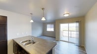This single-floor residence presents an open-concept design that seamlessly connects the kitchen, dining, and living areas. The kitchen is equipped with a double-basin stainless steel sink set into a dark laminate countertop, complemented by elegant pendant lighting that hangs from the smooth white ceiling, adding a stylish focal point to the space. The flooring transitions from durable wood-style planks in the kitchen and living area to plush carpeting in the dining zone, creating distinct yet harmonious areas within the open layout. Natural light fills the space through a window and a full sliding glass door, the latter providing direct access to an outdoor balcony with a railing and open views of the surrounding landscape. The neutral gray walls throughout contribute to a clean, modern aesthetic that pairs well with both the warm and cool tones present in the space. The overall ambiance is bright, airy, and practical, making it an inviting environment for everyday living and entertaining.