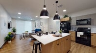 This single-floor unit showcases a thoughtfully designed open-concept layout that seamlessly connects the kitchen and living areas. The kitchen is a standout feature, anchored by a generous island with a crisp white countertop and warm wood cabinetry, illuminated by a pair of striking black pendant lights. Black appliances and a classic subway tile backsplash complement the modern aesthetic, while light wood-tone flooring extends throughout the main living spaces.

The living area receives natural light through large windows fitted with sheer roller shades, creating a bright and welcoming atmosphere. The space flows naturally from the kitchen, offering a comfortable setting for both relaxing and entertaining.

The bedrooms are compact yet functional, each featuring windows that bring in natural light and provide exterior views. Carpeted floors, built-in shelving, and dedicated study areas make these rooms practical and comfortable.

The bathrooms are clean and contemporary in design. One features an open vanity with a white countertop, a wall-mounted open shelf for storage, and wood-look flooring. A second bathroom offers a longer layout with a shower, white vanity, and the same warm flooring, maintaining a cohesive, minimalist aesthetic throughout the unit.