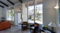 This two-story space showcases a clean, mid-century modern aesthetic defined by its striking architectural details and thoughtful design. The interior is immediately distinguished by expansive floor-to-ceiling windows that span an entire wall, flooding the space with natural light and framing lush outdoor greenery beyond. The large glass panels create a seamless visual connection between the interior and the surrounding landscape, fostering an open, airy atmosphere throughout.

Exposed ceiling beams add architectural character and reinforce the mid-century modern style, contributing to a sense of structured elegance. The dark hardwood flooring grounds the space with warmth and contrast, complementing the neutral white and grey tones of the walls and window frames. Seating areas positioned near the windows invite occupants to enjoy the outdoor views in comfort.

The interior benefits from strong geometric lines and a restrained color palette that keeps the focus on natural materials and light. A glass-paneled entry door further enhances the transparency of the space. Across two floors, the layout offers a thoughtful balance of openness and defined living areas, making this a well-composed space that integrates natural surroundings with refined architectural design.