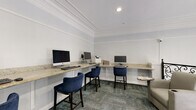 This single-floor shared amenity space combines a functional business center with a refined lounge area, unified by a consistent design aesthetic. Crown molding runs throughout, lending a classic, polished character to the white-painted walls and recessed lighting overhead.

The business center occupies one portion of the space, featuring an L-shaped granite-topped counter that wraps along two walls, providing ample workspace. Multiple desktop computers and a printer are positioned along the counter, complemented by tall navy blue upholstered chairs. Built-in lower shelving beneath the counter adds additional storage and display space. Patterned carpet tiles in muted blue-green tones cover the floor throughout.

Adjacent to the workstation area, a comfortable lounge zone offers plush gray armchairs arranged around a small side table, ideal for casual reading or relaxation. A striking large-scale abstract painting in blue, gold, and green tones serves as a prominent decorative focal point near the entryway. An ornate black wrought iron railing with scrollwork detailing defines the boundary of the upper-level overlook, adding an architectural elegance to the space. A crystal chandelier is visible beyond the railing, hinting at a grander entry area below.
