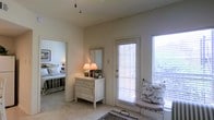 This single-floor space exudes a warm, relaxed atmosphere characterized by soft neutral tones and a cottage-inspired design sensibility. The main living area is finished with plush carpeting underfoot and smooth walls painted in a warm off-white, creating a calming backdrop throughout. Natural light is a defining feature, with large windows fitted with slatted blinds that allow filtered light to illuminate the interior while maintaining privacy. Glimpses of outdoor greenery are visible through the windows, reinforcing a connection to the surrounding natural environment. A glass-paneled door provides an additional source of natural light and potential access to an outdoor area. The space is thoughtfully appointed, featuring a charming whitewashed dresser with a wall-mounted mirror above, accented by soft lamplight for a cozy ambiance. A cushioned accent chair positioned near the window offers a comfortable reading or relaxation nook. An adjacent bedroom, visible through an open doorway, continues the soft, neutral palette with a neatly made bed and warm lighting. The overall layout feels cohesive and inviting, with a quiet, understated charm throughout.