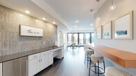 Located at 4555 Minnehaha Ave in Minneapolis's vibrant Longfellow neighborhood, this thoughtfully designed amenity space offers a welcoming and modern environment across a single floor. The open layout connects several distinct functional zones, each finished with contemporary materials and warm, neutral tones throughout.

The lounge area serves as a central gathering point, anchored by a floor-to-ceiling tiled fireplace surround, a plush sofa, and pendant lighting. Large black-framed windows and sliding glass doors flood the space with natural light and provide direct access to a spacious outdoor balcony, where sweeping views of the surrounding Minneapolis greenery and neighborhood skyline can be enjoyed.

The dining area features an oval wood table with seating for multiple guests, illuminated by striking copper pendant lights. A large-scale illustrated mural depicting Minneapolis neighborhoods adds a locally inspired artistic focal point to the room.

A well-appointed wet bar runs along one wall, finished with white cabinetry, a dark countertop, a stainless steel sink, and a distinctive textured tile backsplash. A built-in bar counter with bar stools provides additional seating for casual use. Recessed lighting and globe pendants complete the refined yet functional aesthetic.

The space also includes an ADA-compliant restroom, fully tiled with grab bars and wide clearances. Dark hardwood-style flooring ties the entire space together cohesively.
