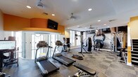 The fitness center at 1670 Kettner in San Diego is a well-appointed, single-floor facility designed to support a comprehensive workout experience. The space features a contemporary layout with a neutral and energetic color palette, highlighted by a bold orange accent wall that adds vibrancy to the environment. Recessed lighting throughout the ceiling, complemented by multiple ceiling fans, ensures the space remains well-lit and comfortable during workouts. A wall-mounted flat-screen television is positioned for easy viewing from cardio equipment. The cardio zone includes multiple treadmills and an elliptical machine, all arranged to face natural light from large windows, creating an open and airy atmosphere. A dedicated boxing area features a punching bag and speed bag setup, while a rack of free weights and additional strength training equipment round out the facility. A large-scale motivational mural depicting a kettlebell adds a dynamic, athletic character to the space. A glass-enclosed area provides additional utility within the footprint. The combination of natural light, modern equipment, and thoughtful design details makes this fitness center a functional and motivating environment for residents.