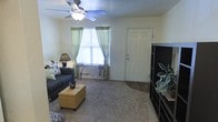This single-floor space features a living room that serves as the central gathering area, finished with wall-to-wall carpeting that adds warmth underfoot. A ceiling fan with an integrated light fixture is mounted overhead, providing both air circulation and ambient lighting. A window dressed with green curtains allows natural light to filter into the room, contributing to a bright and welcoming atmosphere. The front entry door is positioned adjacent to the window, offering a straightforward and accessible layout. The living area accommodates a dark upholstered sofa paired with a tan storage ottoman, creating a functional and comfortable seating arrangement. A black shelving unit along one wall provides display and storage space. The transition between the carpeted living area and what appears to be hard flooring near the entryway adds a subtle distinction between zones. The neutral wall tones keep the space feeling open and versatile, while decorative touches such as throw pillows and a tabletop plant introduce personality. Overall, the space presents a practical and comfortable layout suited for everyday living.