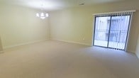 This single-floor space presents a generously sized main living area finished with plush, neutral-toned wall-to-wall carpeting that lends a clean and comfortable feel underfoot. The room's pale yellow walls create a warm, understated backdrop, complemented by crisp white baseboards that frame the perimeter neatly. A classic multi-arm chandelier serves as a focal point overhead, providing both ambient lighting and a touch of traditional elegance to the otherwise understated space.

A sliding glass door fitted with vertical blinds is positioned along one wall, allowing natural light to filter into the room while offering direct access to an outdoor area, likely a balcony or patio. The exterior view hints at a brick building nearby, suggesting a residential setting. The ceiling-mounted HVAC vent indicates a central air system, contributing to the room's functional infrastructure. Overall, the space maintains a simple, neutral aesthetic well-suited for a variety of furnishing styles, making it a versatile and move-in ready environment with a calm, airy ambiance.
