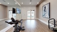 Located on Francis Way in Bloomfield, Connecticut, this single-floor fitness studio offers a well-appointed space designed for movement, cycling, and group exercise. The studio features warm-toned luxury vinyl plank flooring throughout, providing a durable yet visually inviting surface suited for a variety of workout activities. Recessed lighting illuminates the room evenly, creating a bright and energizing environment conducive to exercise.

A prominent floor-to-ceiling mirror spans one full wall, amplifying the sense of space and allowing participants to monitor their form during workouts. Mounted flat-screen televisions are positioned for clear visibility across the room, supporting instructor-led or on-demand fitness programming. A row of stationary spin cycles is arranged to face the mirrored wall, making this an ideal space for cycling classes.

The studio is accessed through a set of double doors with frosted glass panels, contributing to both privacy and a polished, professional appearance. Wall-mounted hooks near the entrance provide convenient storage for personal belongings. A compact equipment rack near the entry offers organized storage for accessories.

Thoughtful decorative touches, including a large framed black-and-white photograph of a ballet dancer, add an artistic element that reinforces the space's dedication to movement and physical discipline. Neutral taupe and white tones throughout create a clean, modern ambiance.