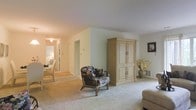 This single-floor residence presents a welcoming, understated interior defined by neutral color tones and wall-to-wall carpeting throughout. The main living area is generously proportioned, offering an open and airy feel enhanced by light-colored walls that create a calm, cohesive atmosphere. A tall, cream-colored armoire serves as a notable furniture piece, providing ample storage while contributing to the room's classic aesthetic. A rattan accent chair with floral upholstery adds a casual, relaxed touch, complemented by a decorative vase of dried florals nearby. A framed piece of wall art punctuates the space without overwhelming the clean lines of the room. A hallway visible from the living area leads to additional spaces, including what appears to be a dining area furnished with a table and chairs, suggesting a functional and connected floor plan. The overall ambiance is quiet and residential, characterized by soft carpeting, subdued lighting, and a straightforward layout that prioritizes comfort and practicality throughout the home.