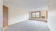 Located at 224 Smith Ave N in Saint Paul, MN, this one-bedroom, one-bathroom unit offers a practical and well-organized layout across a single floor. The generously sized living area is carpeted in light gray and features two wood-framed windows that allow natural light to fill the room while offering views of the surrounding neighborhood. A wall-mounted air conditioning unit and baseboard heating provide year-round climate control. The kitchen is equipped with warm oak cabinetry, a granite-look countertop, a stainless steel sink, a gas range, and a full-size refrigerator, alongside durable wood-look flooring. The spacious primary bedroom also features plush carpeting, warm-toned wood trim, and includes a dedicated closet with built-in shelving for ample storage. A hallway connects the main rooms, providing an efficient flow throughout the unit. The bathroom is thoughtfully finished with white tile wainscoting, a wood-framed medicine cabinet mirror, a white vanity, and a distinctive teal mosaic tile floor, adding a touch of character. Overall, the unit presents a clean, functional living space with consistent warm wood accents and a bright, neutral color palette throughout.