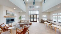This single-floor community clubhouse is designed with a modern farmhouse aesthetic, blending clean lines with warm, inviting materials throughout. The centerpiece of the space is a dramatic great room featuring soaring double-height ceilings, expansive black-framed floor-to-ceiling windows, and French doors that open to an outdoor pool area, flooding the interior with natural light. A sleek linear fireplace with a natural wood mantel anchors the lounge seating area, which is furnished with leather chairs, a plush sofa, and layered textiles that create a comfortable, residential feel. Wood-look tile flooring in a herringbone pattern flows seamlessly through the entry and common areas, transitioning to lighter wood-look planks in the lounge. The kitchen is well-appointed with light gray shaker-style cabinetry, granite countertops, stainless steel appliances, and a large central island illuminated by globe pendant lights. A casual dining and meeting area features bar-height tables with wood and metal stools. The game and activity zone includes a shuffleboard table, wall-mounted television, and additional seating, all beneath the same soaring ceilings. Together, the spaces offer a cohesive, functional, and stylish environment suited for community gatherings and everyday social interaction.