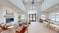 This single-floor community clubhouse is designed with a modern farmhouse aesthetic, blending clean lines with warm, inviting materials throughout. The centerpiece of the space is a dramatic great room featuring soaring double-height ceilings, expansive black-framed floor-to-ceiling windows, and French doors that open to an outdoor pool area, flooding the interior with natural light. A sleek linear fireplace with a natural wood mantel anchors the lounge seating area, which is furnished with leather chairs, a plush sofa, and layered textiles that create a comfortable, residential feel. Wood-look tile flooring in a herringbone pattern flows seamlessly through the entry and common areas, transitioning to lighter wood-look planks in the lounge. The kitchen is well-appointed with light gray shaker-style cabinetry, granite countertops, stainless steel appliances, and a large central island illuminated by globe pendant lights. A casual dining and meeting area features bar-height tables with wood and metal stools. The game and activity zone includes a shuffleboard table, wall-mounted television, and additional seating, all beneath the same soaring ceilings. Together, the spaces offer a cohesive, functional, and stylish environment suited for community gatherings and everyday social interaction.